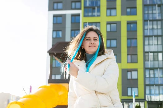 A woman with vibrant blue hair stands confidently in front of a modern building. She gazes ahead, exuding a sense of urban style and individuality.