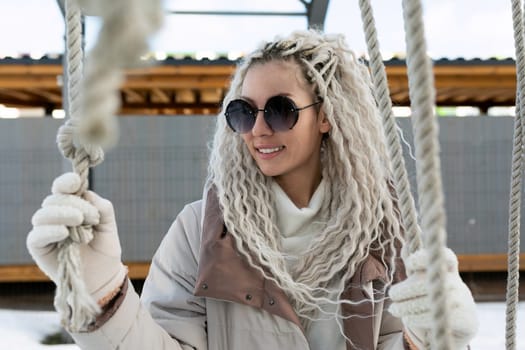 A woman is sitting on a swing, wearing sunglasses and a scarf. She appears relaxed as she swings back and forth gently.