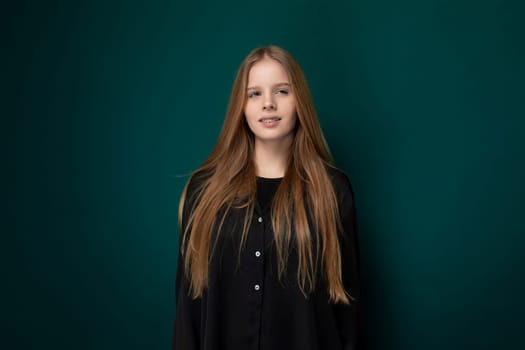 A young girl stands in front of a vibrant green wall, her posture confident and gaze focused. The stark contrast between her figure and the wall creates a striking visual impact, highlighting her presence in the scene.