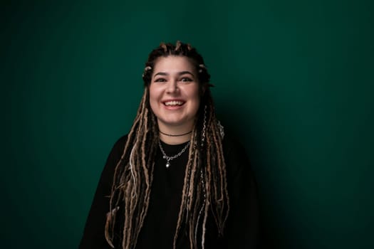 A woman with dreadlocks is standing upright in front of a solid green wall. She appears confident and is looking directly at the camera. The green wall provides a bold background to the scene.