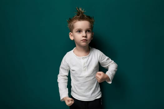 A young male child is standing in front of a bright green painted wall. The boy appears curious as he gazes ahead, with his hands by his sides. The green wall serves as a vibrant backdrop to the scene.