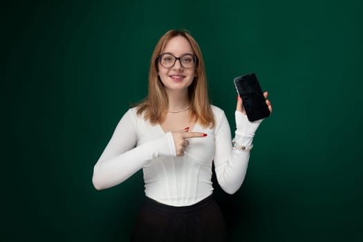 A woman wearing a white shirt is standing, holding a cell phone in her hand. The focus is on her using the phone, with no other visible activities or objects in the background.