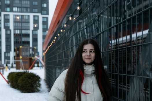 A woman is standing in front of a wooden fence covered in snow. The scene is simple, with the woman looking straight ahead, wearing warm clothing.