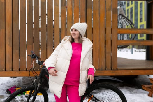 A woman stands next to a bicycle in a snowy landscape, surrounded by white fluffy snow. She is wearing warm winter clothing, looking at the bike.