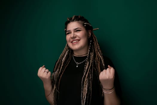 A woman with dreadlocks is standing in front of a bright green wall. She has a confident posture and is looking directly at the camera. The contrast between her hair and the wall creates a striking image.