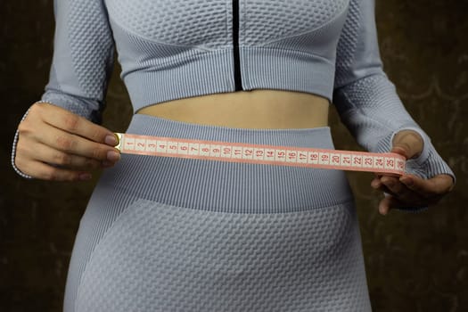 Skinny girl in suit for training at gym holds flexible tape for measuring her waist in hands