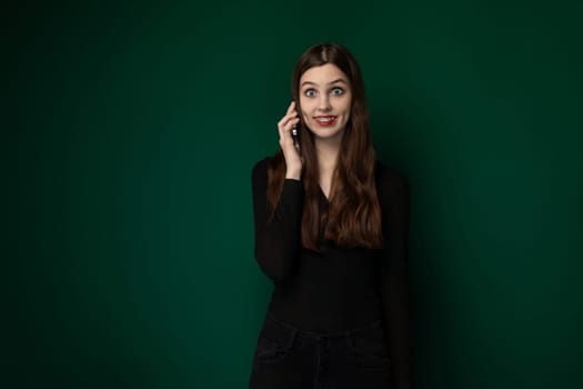 A woman standing in front of a green wall, holding a cell phone to her ear and engaged in conversation. She appears focused on the call, with a neutral expression on her face.