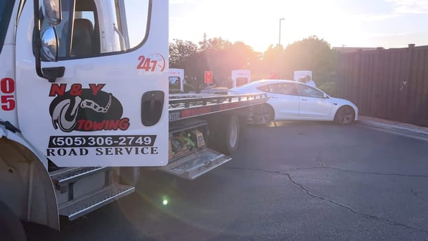 Santa Fe, New Mexico, USA-June 10, 2024-Slow motion-A Tesla electric vehicle is being delivered by a tow truck to a Tesla Supercharger station after running out of charge. The scene captures the car being carefully handled by a worker, who is preparing to connect it for recharging. The Supercharger station is situated in a well-maintained area, with several other charging units visible in the background, indicating a dedicated facility for electric vehicle charging. The image highlights the support and infrastructure available for electric vehicle owners, ensuring they can continue their journey with minimal disruption.