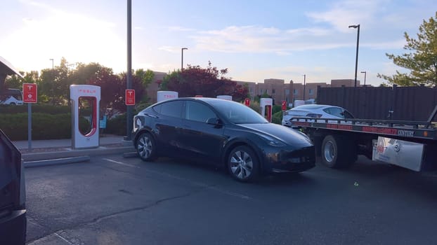 Santa Fe, New Mexico, USA-June 10, 2024-Slow motion-A Tesla electric vehicle is being delivered by a tow truck to a Tesla Supercharger station after running out of charge. The scene captures the car being carefully handled by a worker, who is preparing to connect it for recharging. The Supercharger station is situated in a well-maintained area, with several other charging units visible in the background, indicating a dedicated facility for electric vehicle charging. The image highlights the support and infrastructure available for electric vehicle owners, ensuring they can continue their journey with minimal disruption.