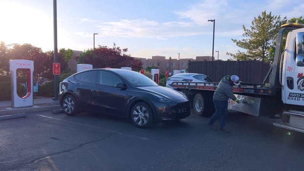 Santa Fe, New Mexico, USA-June 10, 2024-Slow motion-A Tesla electric vehicle is being delivered by a tow truck to a Tesla Supercharger station after running out of charge. The scene captures the car being carefully handled by a worker, who is preparing to connect it for recharging. The Supercharger station is situated in a well-maintained area, with several other charging units visible in the background, indicating a dedicated facility for electric vehicle charging. The image highlights the support and infrastructure available for electric vehicle owners, ensuring they can continue their journey with minimal disruption.