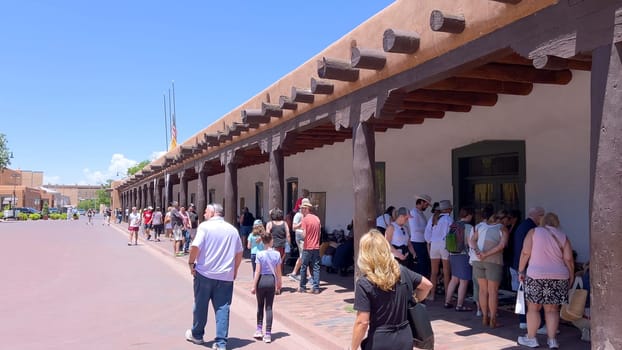 Santa Fe, New Mexico, USA-June 11, 2024-Slow motion-A picturesque covered walkway in Santa Fe, New Mexico, featuring traditional adobe architecture and vibrant blue window frames. The pathway is lined with shops, including a silversmith, showcasing the city rich cultural heritage and lively atmosphere. Potted plants add a touch of greenery to this charming scene.