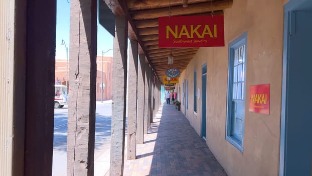 Santa Fe, New Mexico, USA-June 11, 2024-Slow motion-A picturesque covered walkway in Santa Fe, New Mexico, featuring traditional adobe architecture and vibrant blue window frames. The pathway is lined with shops, including a silversmith, showcasing the city rich cultural heritage and lively atmosphere. Potted plants add a touch of greenery to this charming scene.