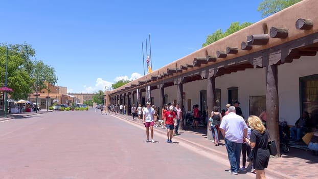 Santa Fe, New Mexico, USA-June 11, 2024-Slow motion-A picturesque covered walkway in Santa Fe, New Mexico, featuring traditional adobe architecture and vibrant blue window frames. The pathway is lined with shops, including a silversmith, showcasing the city rich cultural heritage and lively atmosphere. Potted plants add a touch of greenery to this charming scene.