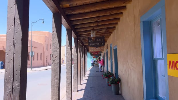 Santa Fe, New Mexico, USA-June 11, 2024-Slow motion-A picturesque covered walkway in Santa Fe, New Mexico, featuring traditional adobe architecture and vibrant blue window frames. The pathway is lined with shops, including a silversmith, showcasing the city rich cultural heritage and lively atmosphere. Potted plants add a touch of greenery to this charming scene.