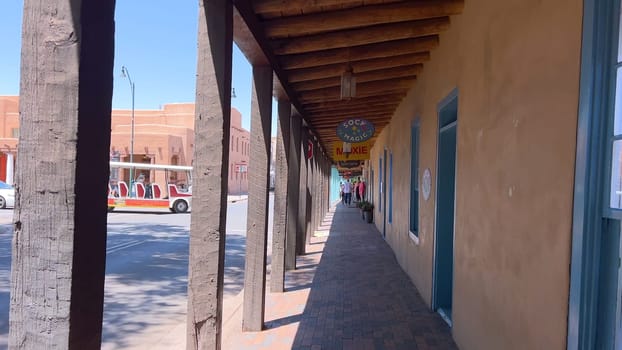 Santa Fe, New Mexico, USA-June 11, 2024-Slow motion-A picturesque covered walkway in Santa Fe, New Mexico, featuring traditional adobe architecture and vibrant blue window frames. The pathway is lined with shops, including a silversmith, showcasing the city rich cultural heritage and lively atmosphere. Potted plants add a touch of greenery to this charming scene.