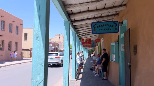 Santa Fe, New Mexico, USA-June 11, 2024-Slow motion-A picturesque covered walkway in Santa Fe, New Mexico, featuring traditional adobe architecture and vibrant blue window frames. The pathway is lined with shops, including a silversmith, showcasing the city rich cultural heritage and lively atmosphere. Potted plants add a touch of greenery to this charming scene.