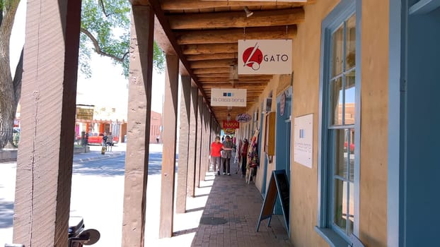 Santa Fe, New Mexico, USA-June 11, 2024-Slow motion-A picturesque covered walkway in Santa Fe, New Mexico, featuring traditional adobe architecture and vibrant blue window frames. The pathway is lined with shops, including a silversmith, showcasing the city rich cultural heritage and lively atmosphere. Potted plants add a touch of greenery to this charming scene.