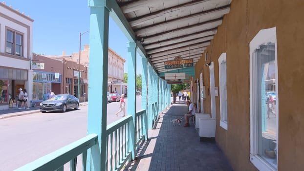 Santa Fe, New Mexico, USA-June 11, 2024-Slow motion-A picturesque covered walkway in Santa Fe, New Mexico, featuring traditional adobe architecture and vibrant blue window frames. The pathway is lined with shops, including a silversmith, showcasing the city rich cultural heritage and lively atmosphere. Potted plants add a touch of greenery to this charming scene.