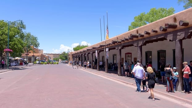 Santa Fe, New Mexico, USA-June 11, 2024-Slow motion-A picturesque covered walkway in Santa Fe, New Mexico, featuring traditional adobe architecture and vibrant blue window frames. The pathway is lined with shops, including a silversmith, showcasing the city rich cultural heritage and lively atmosphere. Potted plants add a touch of greenery to this charming scene.