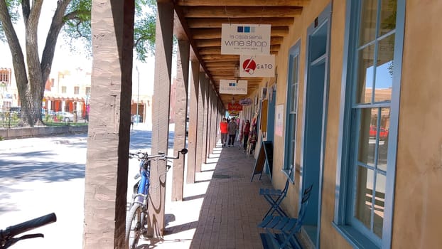 Santa Fe, New Mexico, USA-June 11, 2024-Slow motion-A picturesque covered walkway in Santa Fe, New Mexico, featuring traditional adobe architecture and vibrant blue window frames. The pathway is lined with shops, including a silversmith, showcasing the city rich cultural heritage and lively atmosphere. Potted plants add a touch of greenery to this charming scene.