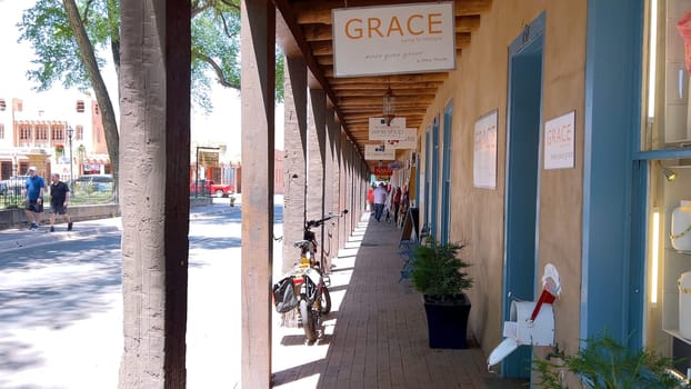 Santa Fe, New Mexico, USA-June 11, 2024-Slow motion-A picturesque covered walkway in Santa Fe, New Mexico, featuring traditional adobe architecture and vibrant blue window frames. The pathway is lined with shops, including a silversmith, showcasing the city rich cultural heritage and lively atmosphere. Potted plants add a touch of greenery to this charming scene.