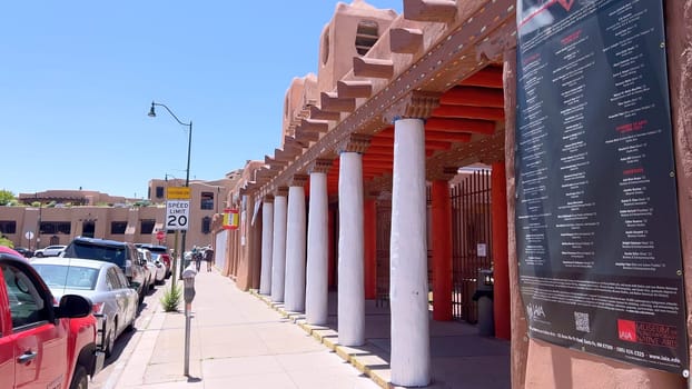 Santa Fe, New Mexico, USA-June 11, 2024-Slow motion-A picturesque covered walkway in Santa Fe, New Mexico, featuring traditional adobe architecture and vibrant blue window frames. The pathway is lined with shops, including a silversmith, showcasing the city rich cultural heritage and lively atmosphere. Potted plants add a touch of greenery to this charming scene.