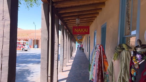 Santa Fe, New Mexico, USA-June 11, 2024-Slow motion-A picturesque covered walkway in Santa Fe, New Mexico, featuring traditional adobe architecture and vibrant blue window frames. The pathway is lined with shops, including a silversmith, showcasing the city rich cultural heritage and lively atmosphere. Potted plants add a touch of greenery to this charming scene.