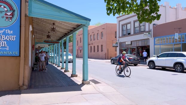 Santa Fe, New Mexico, USA-June 11, 2024-Slow motion-A picturesque covered walkway in Santa Fe, New Mexico, featuring traditional adobe architecture and vibrant blue window frames. The pathway is lined with shops, including a silversmith, showcasing the city rich cultural heritage and lively atmosphere. Potted plants add a touch of greenery to this charming scene.