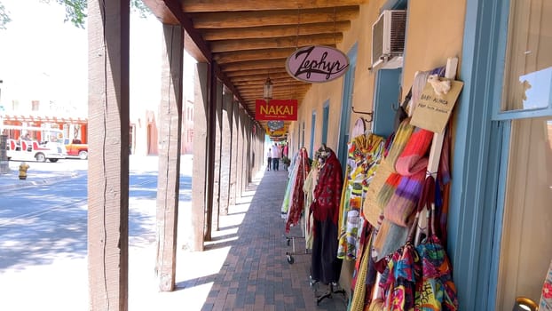 Santa Fe, New Mexico, USA-June 11, 2024-Slow motion-A picturesque covered walkway in Santa Fe, New Mexico, featuring traditional adobe architecture and vibrant blue window frames. The pathway is lined with shops, including a silversmith, showcasing the city rich cultural heritage and lively atmosphere. Potted plants add a touch of greenery to this charming scene.