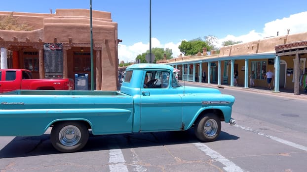 Santa Fe, New Mexico, USA-June 11, 2024-Slow motion-A vintage blue pickup truck drives through a historic downtown area, showcasing charming adobe buildings and a vibrant atmosphere on a sunny day.