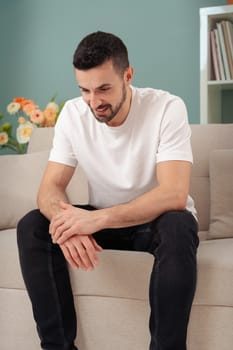 Portrait of a handsome and tired young man in casual clothes looking down while sitting at home. Depressed bearded man. Attractive stressed businessman. Business People Concept.