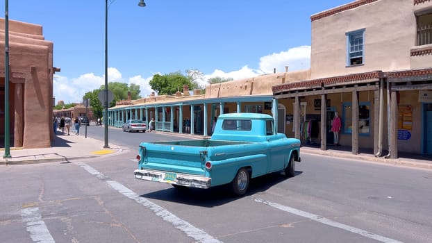 Santa Fe, New Mexico, USA-June 11, 2024-Slow motion-A vintage blue pickup truck drives through a historic downtown area, showcasing charming adobe buildings and a vibrant atmosphere on a sunny day.