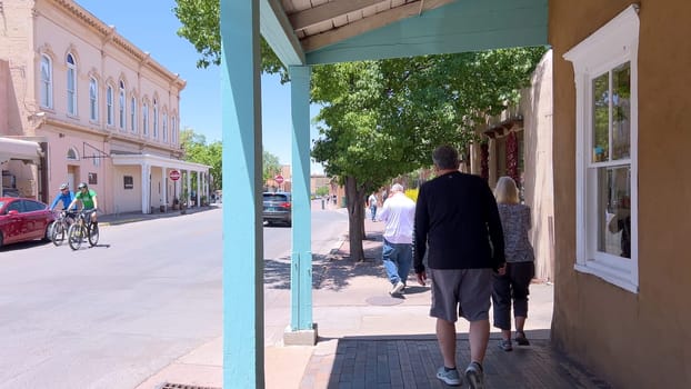 Santa Fe, New Mexico, USA-June 11, 2024-Slow motion-A picturesque covered walkway in Santa Fe, New Mexico, featuring traditional adobe architecture and vibrant blue window frames. The pathway is lined with shops, including a silversmith, showcasing the city rich cultural heritage and lively atmosphere. Potted plants add a touch of greenery to this charming scene.