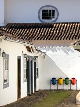 Brightly colored waste bins are lined up beside a white-painted house with terracotta roof tiles.