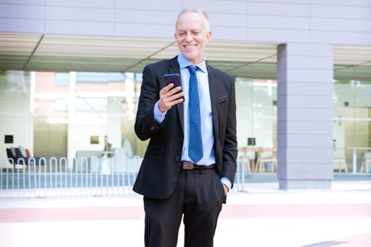 Satisfied with the results, the businessman walks on the street outside the office building, a mature boss holds a phone in his hands, writes messages and reads news online, using an application.