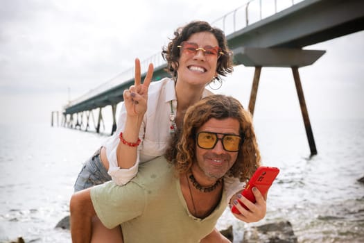 Happy caucasian man carrying his girlfriend on his back with smartphone, having fun together on the beach.