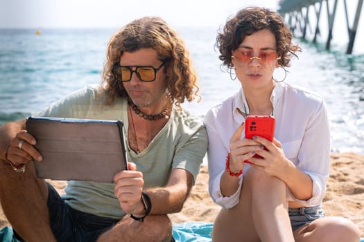 Middle-aged man and woman sitting on the beach with surprised face, browsing smartphone apps. looking at camera Concept:Holidays y Technology