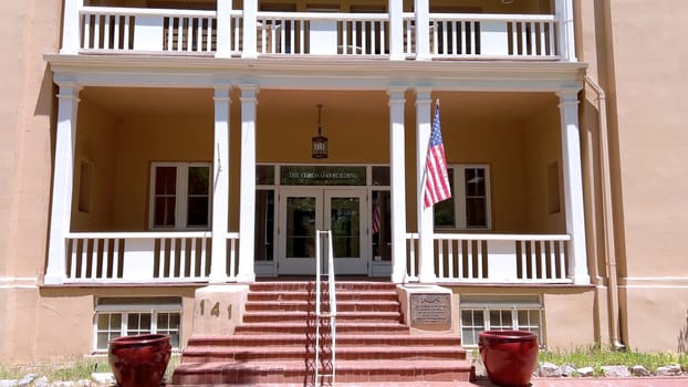 Santa Fe, New Mexico, USA-June 10, 2024-Slow motion-A historic building in a downtown area featuring a symmetrical facade with two levels of balconies, an American flag, and well-maintained landscaping on a sunny day.