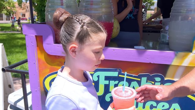 Santa Fe, New Mexico, USA-June 11, 2024-Slow motion-A young girl with a stylish braided bun hairstyle receiving a refreshing fruit juice at a vibrant and colorful stand. The stand is decorated with bright signs and large beverage containers, indicating a lively atmosphere on a sunny day.