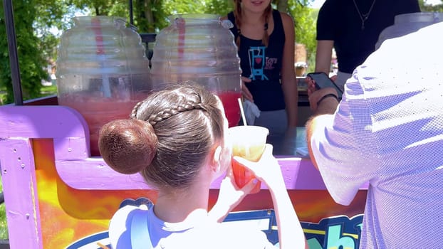 Santa Fe, New Mexico, USA-June 11, 2024-Slow motion-A young girl with a stylish braided bun hairstyle receiving a refreshing fruit juice at a vibrant and colorful stand. The stand is decorated with bright signs and large beverage containers, indicating a lively atmosphere on a sunny day.