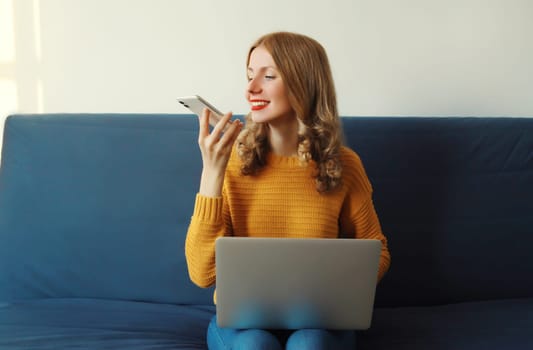 Modern young woman working with laptop talking on phone, takes calling or using voice assistant, command recorder sitting on a couch in living room at home