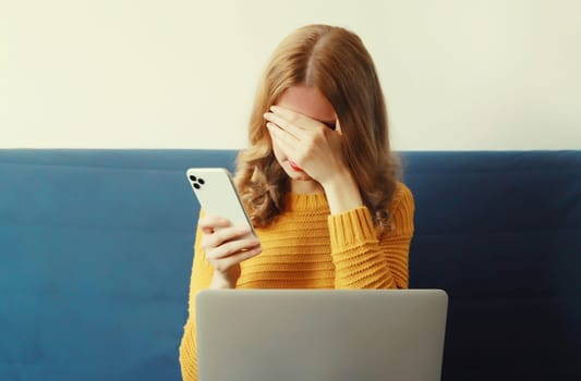 Sad upset crying woman holding phone suffering from headache, migraine while working with laptop sitting on sofa in room at home
