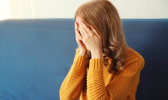 Upset sad crying woman covering her eyes and face with her hands while experiencing mental or physical suffering, fatigue sitting on the couch in room at home