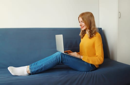 Happy modern young woman working on a laptop, freelancer girl looking at computer sitting on a couch in the living room at home