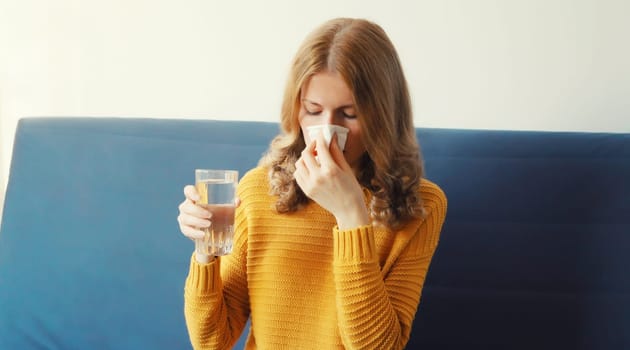 Sick exhausted young woman sneezing blow nose using tissue drinks water from a glass taking a medication at home