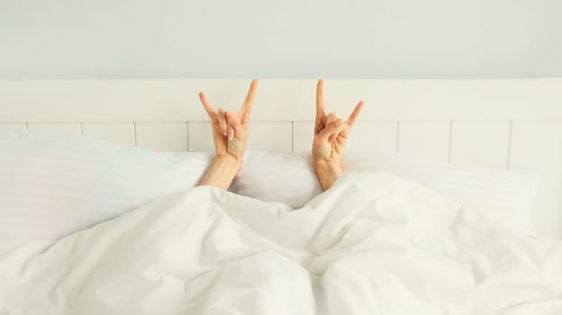 Cheerful lazy woman waking up after sleeping lying in soft comfortable bed showing gesture stretching her hands up from under the blanket in white bedroom