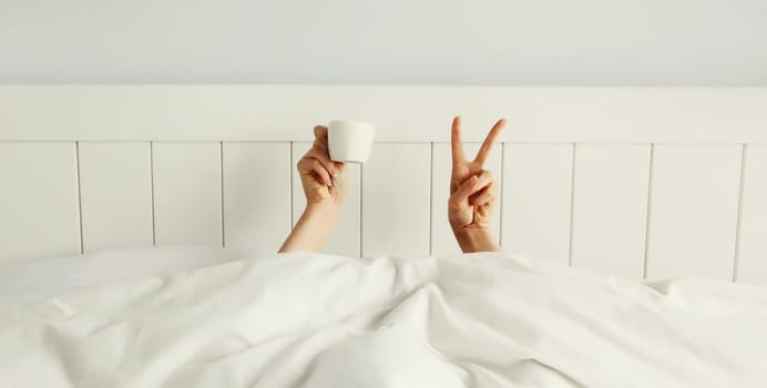 Cheerful lazy woman waking up after sleeping lying in soft comfortable bed showing empty cup coffee stretching her hands up from under the blanket in white bedroom