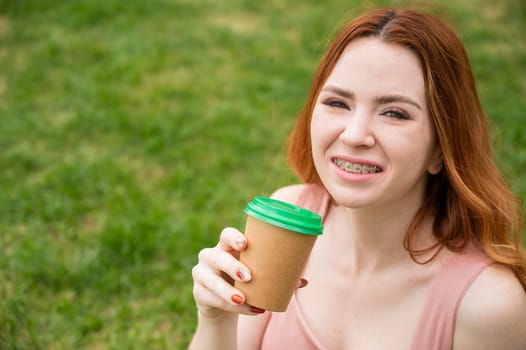 Beautiful young woman with braces on her teeth drinks from a cardboard cup while sitting on the grass