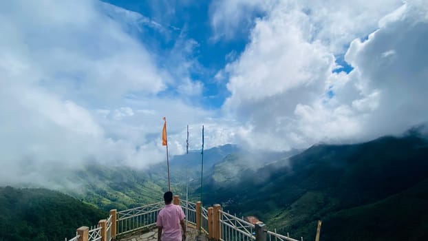 This photo depicts a man standing on a viewing platform overlooking a mountain range shrouded in fog. The platform is made of wood and metal and has a railing. The man is wearing a pink shirt and is looking out at the view. In the background, there are mountains covered in clouds, and the sky is a bright blue.