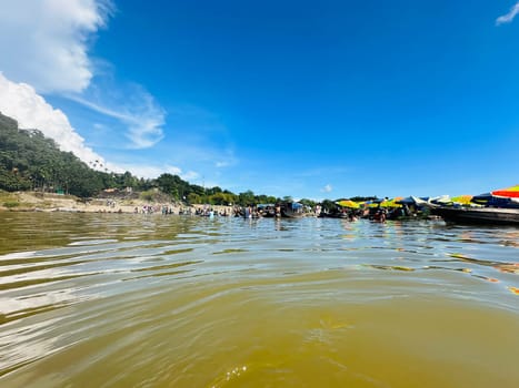 A beautiful view of a tropical river with people swimming and relaxing on the shore. The water is calm and the sky is clear. The scene is filled with colorful umbrellas and boats, giving it a vibrant atmosphere.